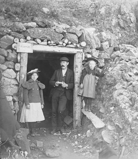 Black and white image. Mr. Hartshorn, Hazel Hartshorn and another child stand in the entrance of a quartz mining tunnel that is surrounded by boulders., Florence M. Hartshorn photographs, 1899-1900