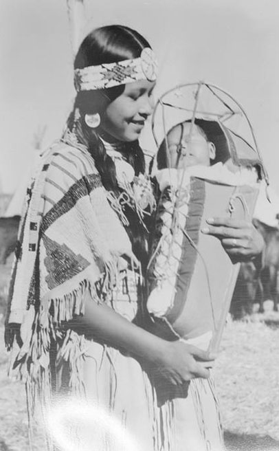A smiling woman in Native American attire holds a sleeping baby that is secured in a cradleboard., Lee D. Drake (1882-1957) photographs, ca. 1910-1957