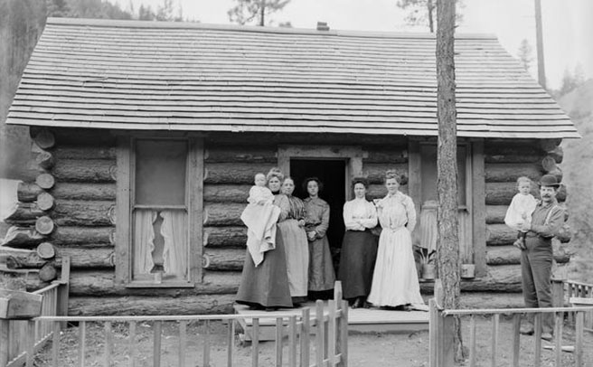 An outside photograph of a group of people&mdash;an adult male holding a baby, and five adult or teenage women, one woman holding a baby&mdash;standing in front of a log cabin., Lee Moorhouse (1850-1926) photographs, 1888-1916