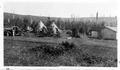 Tipi style tents, man & woman beside truck, cabin, shelter, low bush in foreground, higher elevation in background