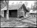 Man sitting in front of log cabin, identified as X. G. Talcott homestead. Tools leaning against doorway.