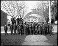 Formal portrait of students at Chemawa Indian School, Chemawa, OR., standing in uniform beneath archway. Trees leading to background building.