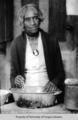Elderly African American woman, wearing white dress, dark sweater, and cameo brooch, standing over a mixing bowl.