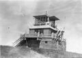 Three story fire lookout made of cut logs. Outside staircases leading up to top floor, 3rd floor has glass windows all around. American flag flying.