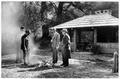 Four men standing around fire with shelter in background with running water pipe