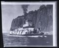 The 'River Queen' sternwheeler on river, tall rock cliffs in background; 2 covered wagons on deck.