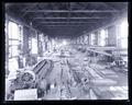 Interior of Factory. Large metal plates are lined up on floor. Machines and workers line walls.