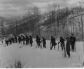 Snow on hillside, bare trees in background.  15 men in a line with walking sticks are walking across a snowy field.  Behind the men are some tents.