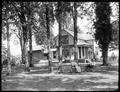 Couple sitting in yard of farmhouse, Story, OR. House in background, surrounded by trees. Woman in chair holding rabbit, hammock behind her.