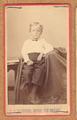 Small boy sitting, wearing trousers and a white shirt with dark tie