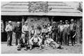 Men standing and seated in front of community kitchen. Kitchen has large stone chimney.