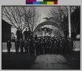 Formal portrait of students at Chemawa Indian School, Chemawa, OR., standing in uniform beneath archway. Trees leading to background building. (recto)