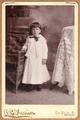 Little girl wearing white outfit with a dark bow, standing by a wicker chair