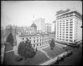 View from above of Post Office and grounds, Portland. Office buildings in background, building under construction across from Post Office. Autos parked along 5th St., in foreground.