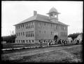 School building at Carlton, with group of students in front of building. Dirt road in foreground.