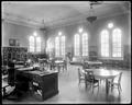 Interior of Albina Library, Portland. Chairs around round tables in room, with desk in foreground. Tables, bookshelves in background, windows above.