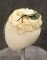 Halo-style hat of a circle of ivory fabric flowers of synthetic and velvet with a corsage at crown with green leaves and brown cord branch