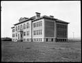 Stone and brick high school building, Story, OR., with large woodpile in background.