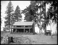 Large log cabin with stone chimney, at Story, OR. Auto parked beside cabin. Chairs on covered porch of cabin.