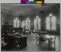 Interior of Albina Library, Portland. Chairs around round tables in room, with desk in foreground. Tables, bookshelves in background, windows above. (recto)