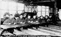 Men with pens and paper sitting at long tables.