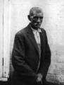 Elderly African American man, wearing overalls, button-up shirt, and suit coat, with white-washed brick wall in background.